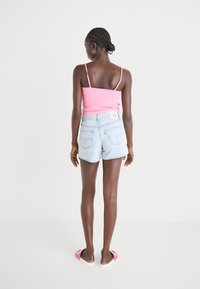 Tirantes delgados en una camiseta de tirantes de color rosa con canalé, combinada con pantalones cortos de denim azul claro. La modelo lleva sandalias rosas sin talón, vista desde la parte de atrás.