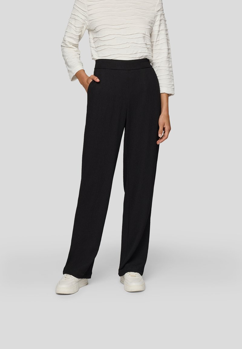 Frau trägt schwarze, hoch taillierte, weit geschnittene Hose, weißes, strukturiertes Langarmshirt und weiße Turnschuhe, steht mit einer Hand in der Tasche.