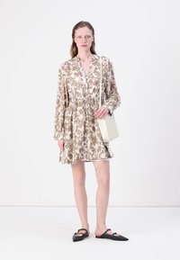 Abito floreale in tonalità beige e marrone, con bottoni e vita regolabile, maniche lunghe. La modella tiene una borsa piccola e indossa scarpe basse nere.