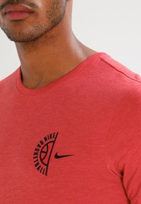 Mężczyzna w czerwonym podkoszulku koszykarskim Nike z czarnym logo i swooshem, ukazujący dolną część twarzy, szyję i górną część klatki piersiowej.