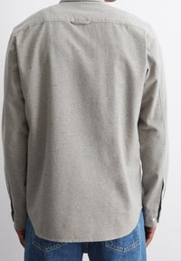 Person, die ein langärmeliges, hellgraues, gesprenkeltes Hemd und blaue Jeans aus Denim trägt, gezeigt von hinten vor einem schlichten Hintergrund.