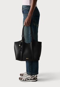 Schwarze Leder-Tote mit strukturiertem Finish, ausgestattet mit zwei Griffen, einem dekorativen Knoten und einer goldenen Logo-Plakette. Trägt man zu verwaschenen blauen Jeans und Sneakers.