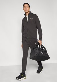 Αθλητικό σκούρο γκρι μπουφάν με φερμουάρ και αντίστοιχο παντελόνι, μαύρα αθλητικά παπούτσια με πράσινες λεπτομέρειες, κρατώντας μαύρη τσάντα γυμναστηρίου με λευκή ραφή.