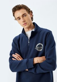 Sudadera de color azul marino con cuello con cremallera, que presenta un parche con un logo circular en el pecho y puños acanalados. Textura suave.
