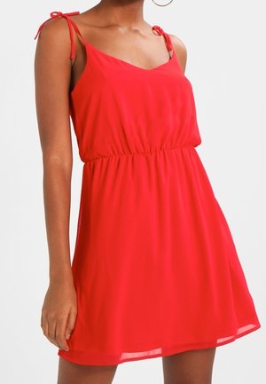 Femme portant une robe rouge sans manches avec des bretelles nouées et une taille élastique, se tenant devant un fond blanc uni.