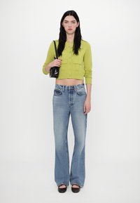 Lime green cropped cardigan with two front pockets, paired with light blue flared jeans and black open-toe shoes. Model holding a small black bag.