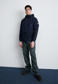 Navy hooded jacket with front pockets, drawstring waist; paired with dark green patterned pants and black clogs with yellow accents.
