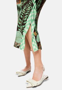 Slingback-Flats aus cremfarbenem Leder mit spitzem Zehenbereich und Schleifenakzent. Der Rock weist ein grünes Karomuster und einen tropischen Blätterdruck auf dunklem Hintergrund auf.