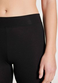 Zwarte legging met een gladde textuur en een brede elastische tailleband, vergezeld door een hand die in de buurt rust, wat een gemiddelde hoogte van de taille laat zien.