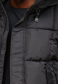 Sort dunjakke med lynlås, justerbar hætte med snore, og syninger for isolering; blød, quiltet tekstur.