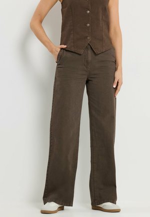 Osoba ubrana w ciemnobrązową kamizelkę zapinaną na guziki oraz pasujące szerokie spodnie, stoi z jedną ręką w kieszeni i białymi butami na nogach.