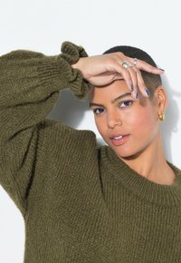 Olivgrüner Strickpullover mit grober Textur und gerippten Bündchen, getragen von einer Person mit erhobener Hand vor einem weißen Hintergrund.