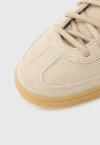 Beżowy zamszowy sneaker z teksturowaną powierzchnią, okrągłym noskiem, tonowymi sznurowadłami oraz gumową podeszwą z wzorzystym bieżnikiem.