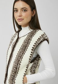 Veste en polaire crème avec motifs géométriques marron, dotée d'un col plié et de côtés ouverts, portée par-dessus un haut à manches longues blanc.