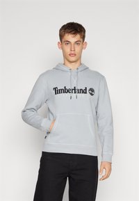 Felpa grigia chiaro con cappuccio regolabile, tasca a marsupio e polsini a costine. Presenta il logo "Timberland" e l'anno di fondazione ricamato in nero.