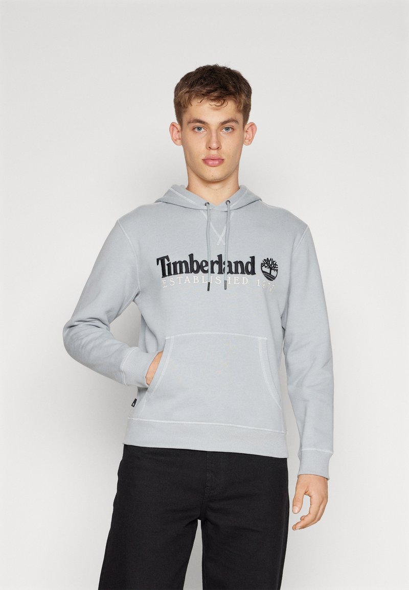 Felpa grigia chiaro con cappuccio regolabile, tasca a marsupio e polsini a costine. Presenta il logo "Timberland" e l'anno di fondazione ricamato in nero.