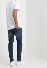 Man med vit t-shirt, mörkblå jeans och vita Reebok-sneakers som står framför en enkel vit bakgrund.