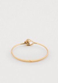 Βραχιόλι από μέταλλο χρυσού χρώματος με λεία, στρογγυλή μπανέλα και διακοσμητικό σε σχήμα καρδιάς στο κλείσιμο. Απλός και κομψός σχεδιασμός.