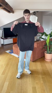 Musta lyhythihainen paita napilla, esillä henkilön päällä. Huoneessa on puulattia ja ruskea sohva, taustalla kasveja.