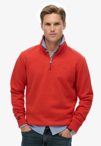 Sudadera de forro polar roja con cuello con cremallera y camisa de botones azul por debajo. Presenta mangas largas y un pequeño logo en el pecho.