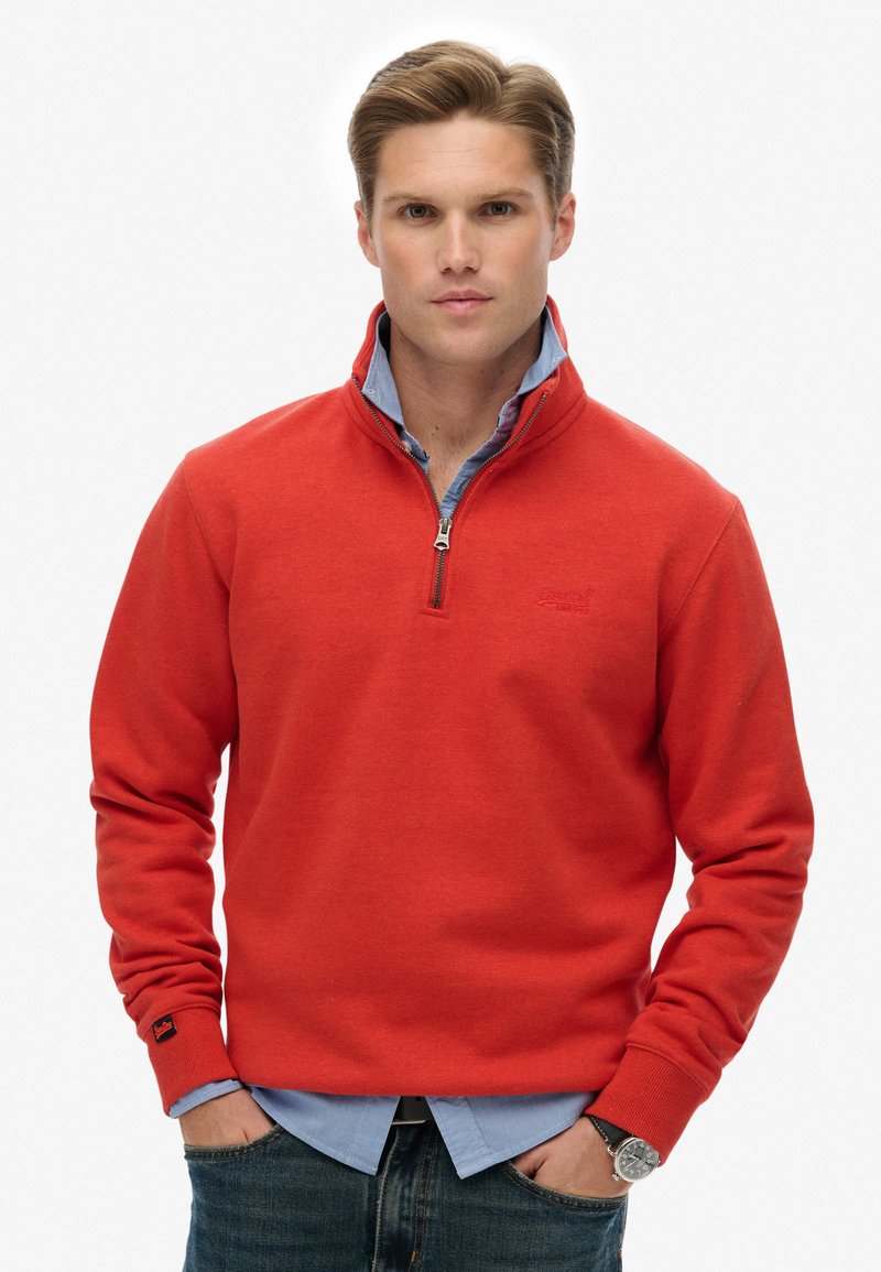 Sudadera de forro polar roja con cuello con cremallera y camisa de botones azul por debajo. Presenta mangas largas y un pequeño logo en el pecho.