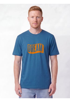 Homme aux cheveux roux courts portant un t-shirt bleu avec un texte orange "DREAM", se tenant devant un fond blanc uni.