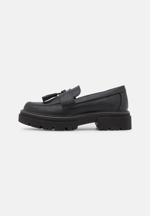 Mocassin en cuir noir avec semelle crantée épaisse et pompon décoratif sur le dessus, présenté de profil droit sur fond uni.