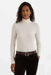 Un haut à col roulé crémeux à manches longues en tissu doux, porté avec un pantalon en velours côtelé marron foncé et une ceinture marron avec une boucle en argent.
