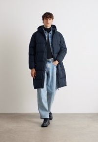 Young man walking in black hooded long puffer coat, black turtleneck, light blue jeans, and black boots against plain white background.