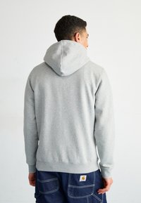 Felpa grigia con cappuccio regolabile, polsini e orlo a coste, e una texture liscia. Indossata con pantaloni cargo blu scuro con cuciture visibili.