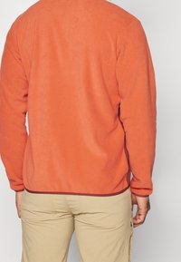 Pullover in pile arancione da uomo con maniche lunghe, tessuto testurizzato e dettaglio del margine bordeaux, abbinato a pantaloni cargo beige chiaro.