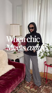 Mujer con sudadera negra con capucha, gafas de sol, pantalones grises y sandalias posa en el interior cerca de un sofá de terciopelo rojo, una mesa auxiliar, una lámpara y una planta en maceta.