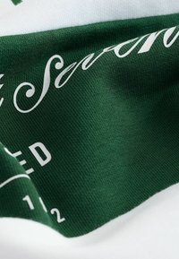 Gros plan sur un tissu vert avec des textes cursifs blancs et en majuscules, montrant un matériau tricoté texturé et des mots et chiffres imprimés partiellement.