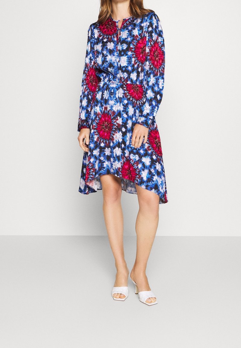 Femme portant une robe à manches longues bleue et blanche à motifs floraux avec de grandes fleurs rouges et des talons blancs à bout ouvert.