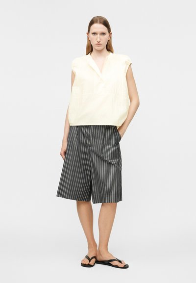 Γυναίκα με αμάνικο κρεμ μπλουζάκι, μαύρο-άσπρο σορτς με ρίγες μέχρι το γόνατο και μαύρα σαγιονάρες, στέκεται με το ένα χέρι στην τσέπη.