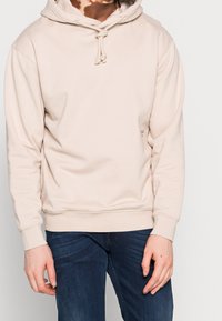 Persona che indossa un hoodie beige chiaro con lacci e jeans blu scuri, in piedi contro uno sfondo chiaro e semplice.