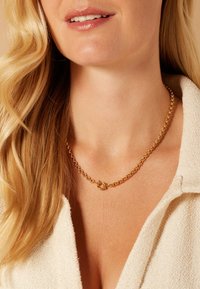 Gouden ketting met onderling verbonden schakels, met een ronde sluiting, gedragen tegen een lichtgekleurde stoffen achtergrond.