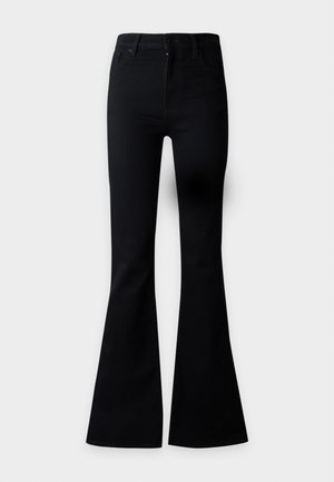Femme aux longs cheveux foncés portant une veste courte noire, un top en maille beige, un pantalon évasé noir et des chaussures pointues beiges sur un fond uni.