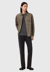 Giacca grigia leggera con due tasche sul petto, chiusura a bottoni, indossata sopra una maglietta scura. Pantaloni neri e stivali neri completano l'outfit.