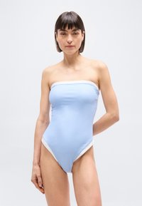 Γυναίκα με κοντά σκούρα μαλλιά που φοράει ένα μπλε ανοιχτό ολόσωμο μαγιό χωρίς τιράντες με λευκή επένδυση, στέκεται μπροστά σε ένα απλό ανοιχτό φόντο.