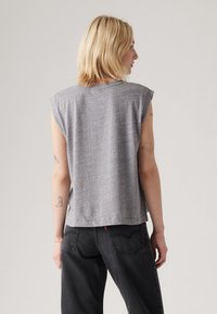 Blonde person facing away wearing a sleeveless gray top and black jeans, with small tattoos on both arms against a plain background.