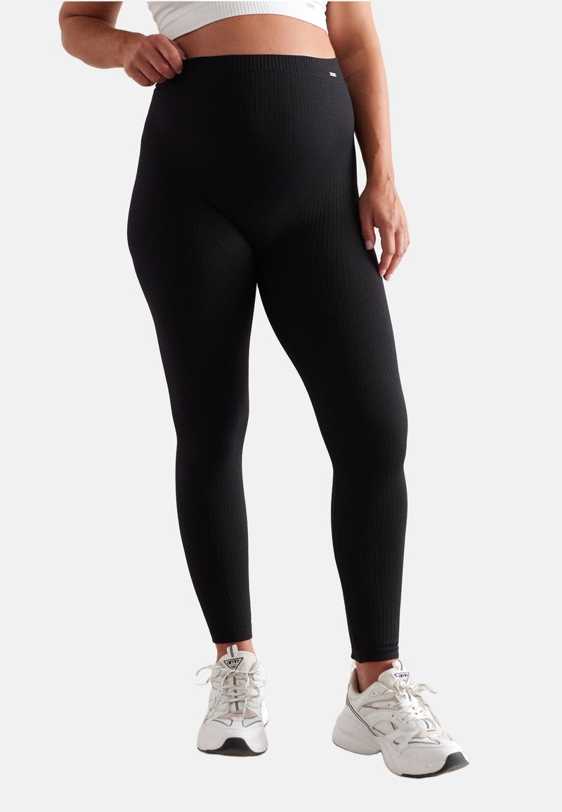 Femme portant des leggings noirs côtelés et des baskets blanches, un top blanc court, debout devant un fond uni.