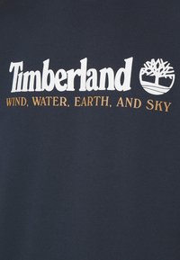 Laivastonsininen paita, jossa valkoinen "Timberland"-logo ja kultaiset sanat "WIND, WATER, EARTH, AND SKY" alla. Sileä kangaspinta.