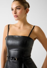 Robe en cuir noir avec un décolleté carré, des bretelles fines, un corsage ajusté, et une taille ceinturée ornée d'un accent doré. Surface texturée.