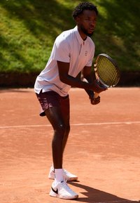 Tennisimängija helesinisest polo särgis ja marraskortsides, hoides racketit. Valged Nike kingad mustade aktsentidega, punasel savi väljakul.