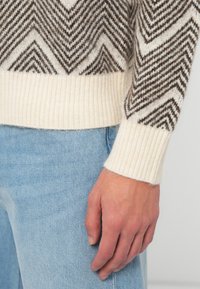 Vuna pulover s chevron uzorkom u smeđoj i krem boji, s rebrastim kremastim obrubom i manžetama. Nosite ga s svijetloplavim trapericama.