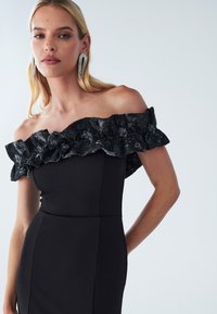 Black dress with off-shoulder design, featuring a textured black ruffled accent. Smooth fabric, figure-hugging silhouette, and minimal seams.