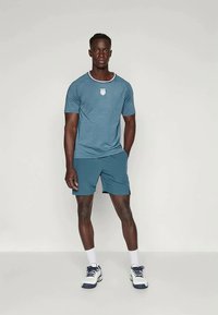 T Türkiz sportpóló, kerek nyakkal és textúrált anyagból, passzoló sorttal párosítva. A pólón középen elhelyezkedő fehér logó. Fehér sportcipő.