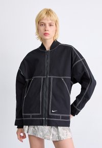 Casaco preto de grande dimensão com costura branca, fecho frontal com zíper e bolsos. O tecido parece texturizado; apresenta um logótipo da Nike na bainha.