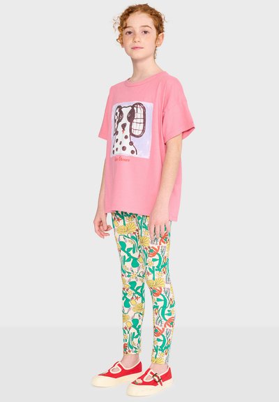 Young person with curly red hair wearing a pink dog-print t-shirt, colorful floral leggings, and red canvas shoes with a buckle.